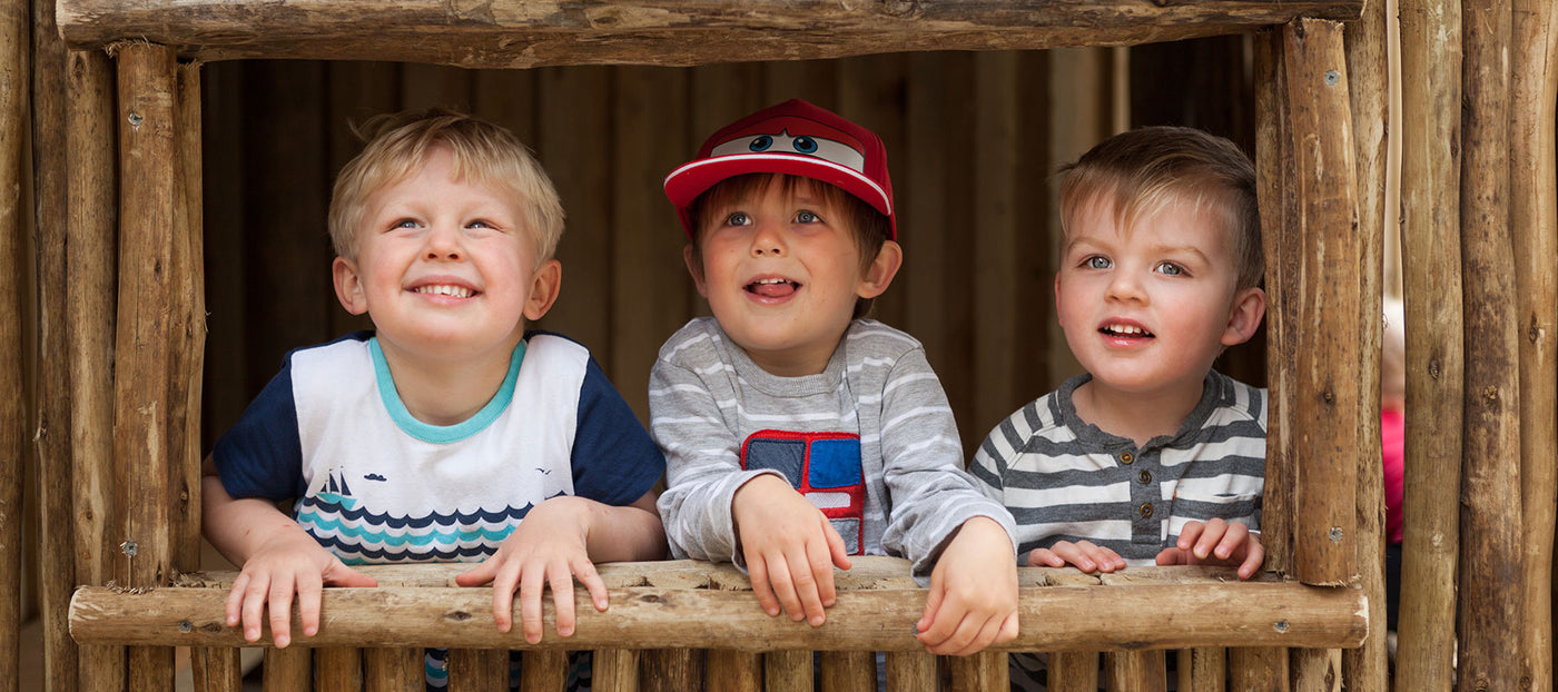 3 preschool boys smiling from a wooden play house at A pot full of colourful felt tip pens at Tadpoles Early Childhood Centre